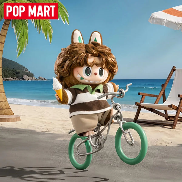 A Luluby vinyl plush pendant character with brown fur and green accents riding a bicycle on a beach.