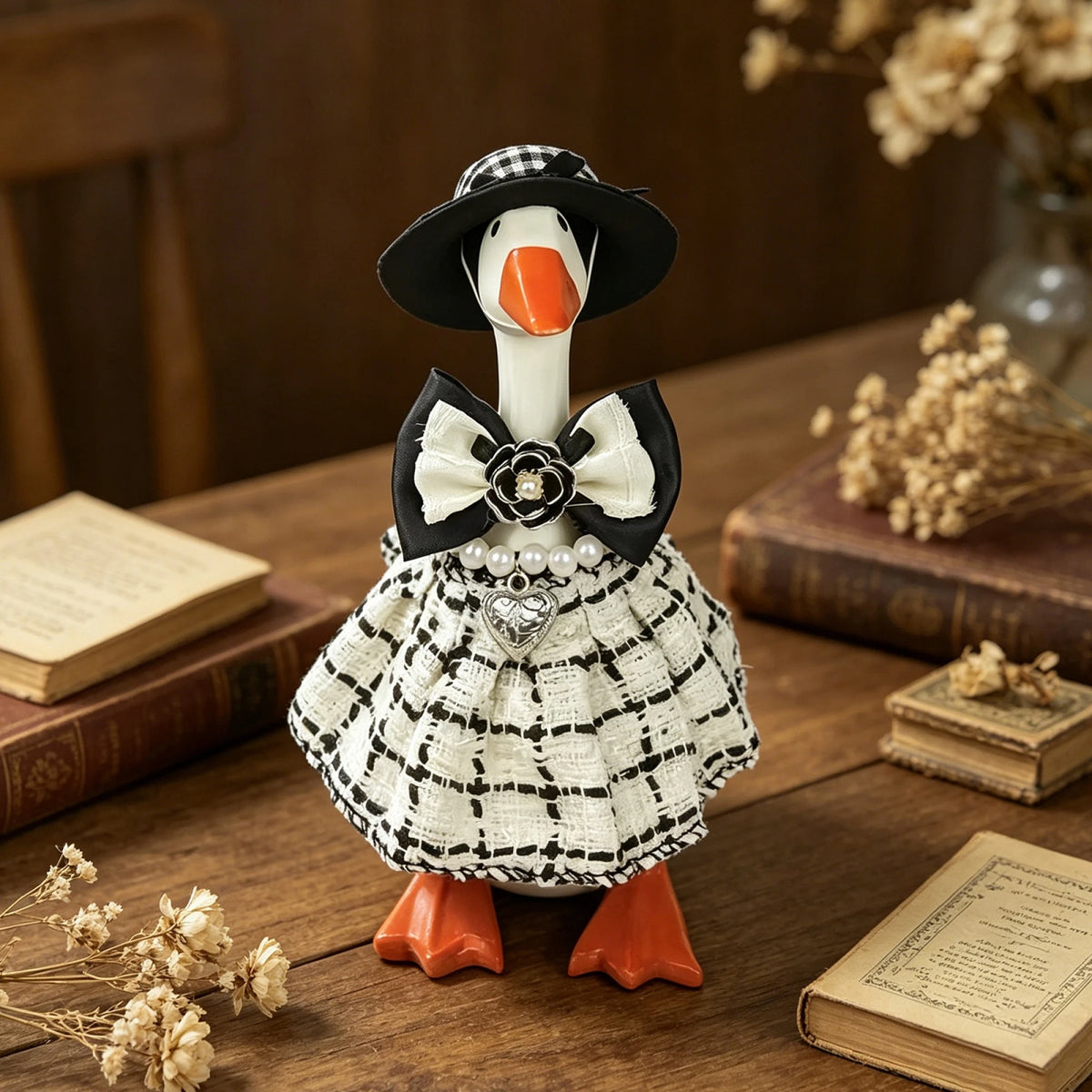 A white goose statue dressed in a black plaid dress and bow, sitting on a wooden surface with books.