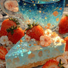 A glass of sparkling blue drink sits next to a slice of strawberry cake decorated with floral elements and fresh strawberries
