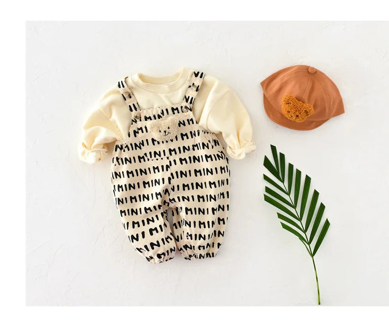 A cream and black striped baby romper with a bear decoration lying next to an orange hat and a green leaf.