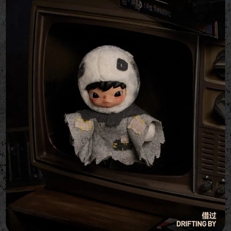 A plush doll wearing a white hood and dark features is displayed inside a wooden box.