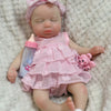 A realistic reborn baby doll with pink hair and a pink ruffled dress lying on a white fluffy surface.