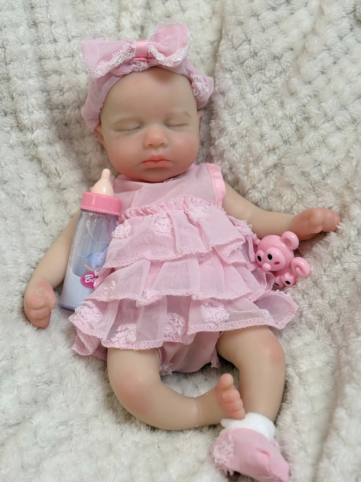 A realistic reborn baby doll with pink hair and a pink ruffled dress lying on a white fluffy surface.