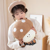 A woman is holding a round, brown mushroom plush toy with white spots against a light background.