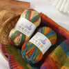 A bundle of three skeins of gradient acrylic yarn in various colors resting in a woven basket and an orange knitted fabric.
