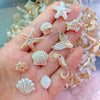 A hand displays a collection of starfish, seashells, and a small fish acrylic bead mix for DIY jewelry making.