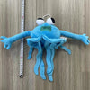 Blaues Plüschtier, das wie eine Quallenfigur mit Tentakeln und grünem Mund aussieht, vor einem Holz-Hintergrund.