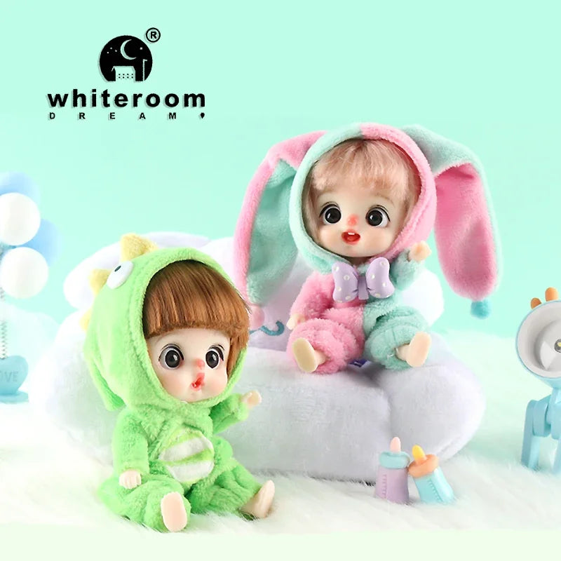 A small, pink and green doll with long ears is shown sitting on white fluff, next to a small blue item.