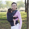 A woman wearing a pink long-sleeved shirt is holding a baby in an adjustable cotton carrier wrap outdoors.