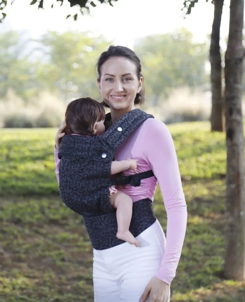 A woman wearing a pink long-sleeved shirt is holding a baby in an adjustable cotton carrier wrap outdoors.