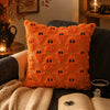 An orange textured pillow with a pattern of black ghost faces, resting on a dark gray sofa next to a knitted blanket and an o