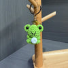 A bright green crochet frog charm with a white belly is hanging from a gold-colored key ring on a wooden display stand.
