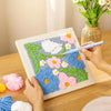 Hands are assembling a flower pattern punch needle embroidery kit featuring various colored wool fibers and floral designs.
