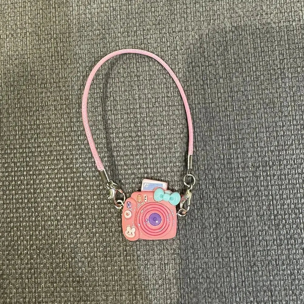 A pink beaded necklace with a small camera charm and a light pink cord against a dark gray fabric background.