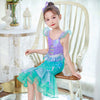 A young girl wearing a purple and teal mermaid fantasy dress with a ruffled fishtail skirt, sitting in a white wooden chair.
