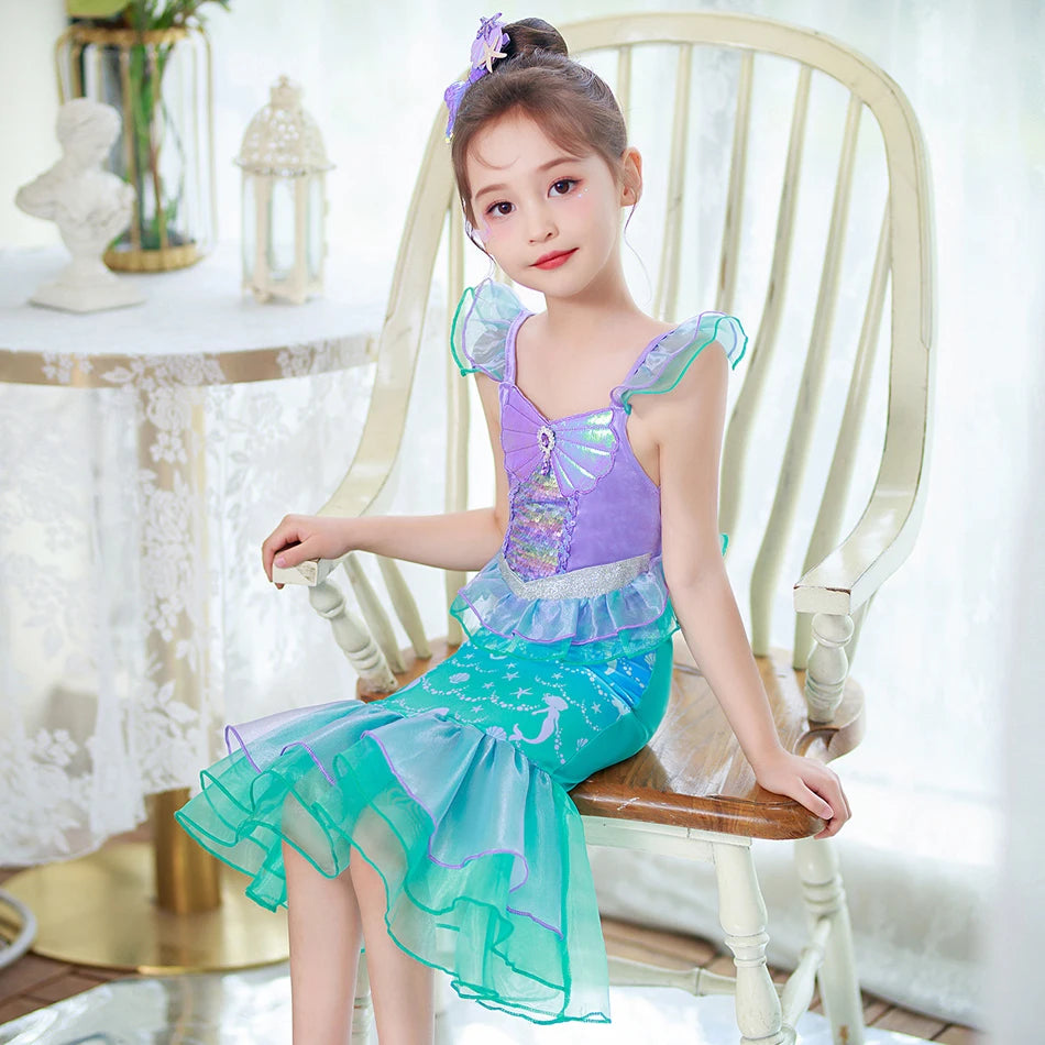 A young girl wearing a purple and teal mermaid fantasy dress with a ruffled fishtail skirt, sitting in a white wooden chair.