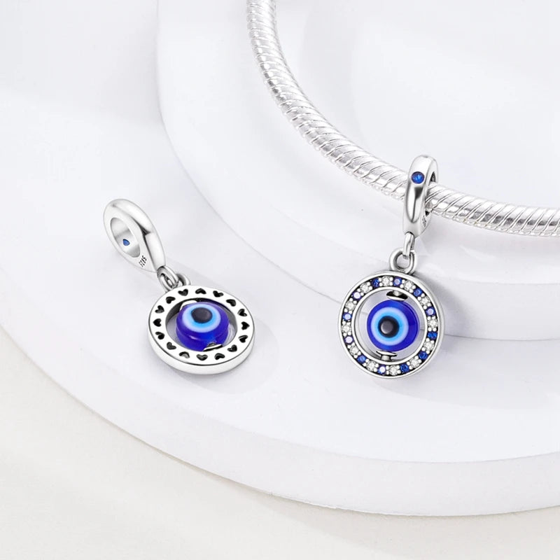 A pair of sterling silver charm beads featuring cross, angel, and evil eye designs displayed on a white surface.