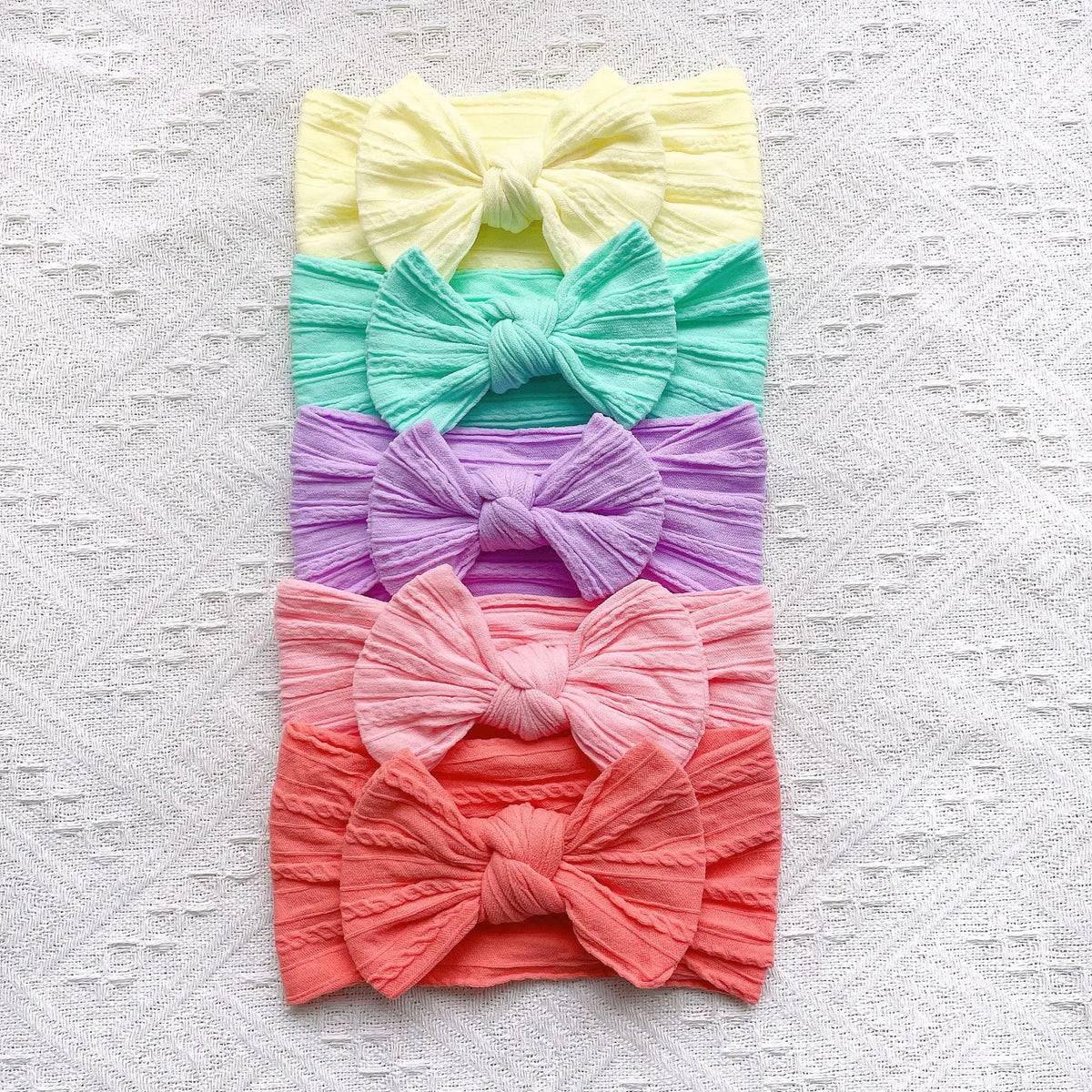 A stack of five pastel colored fabric bow baby headbands in yellow, teal, purple, pink, and coral.