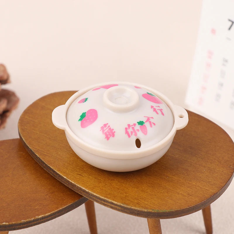 A white miniature copper soup pot with pink decorations sits on a wooden table.