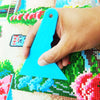 A hand holds a bright blue diamond painting precision tool against a colorful fabric background.