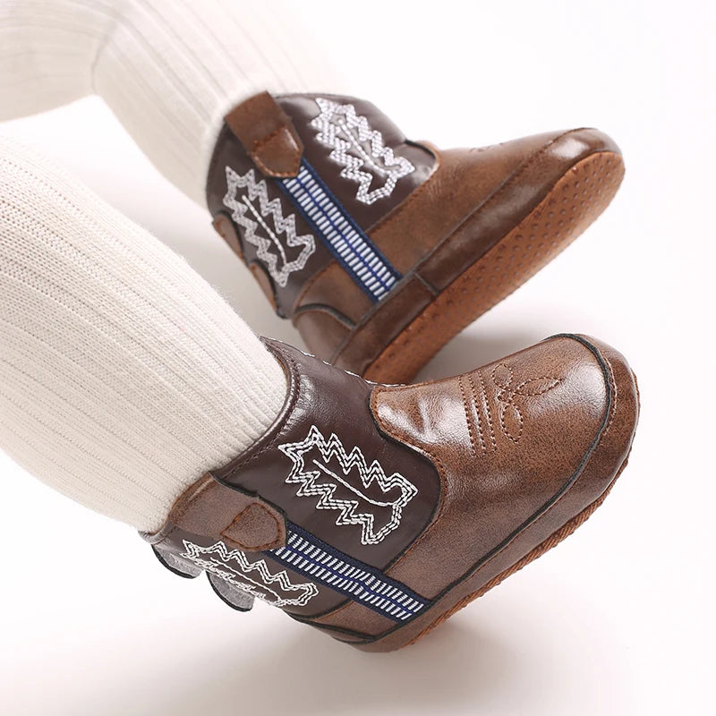 A close-up of brown baby western booties with white embroidery and blue stitching on a white sock.