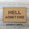 Un écusson rectangulaire beige tissé avec du texte noir indiquant HELL ADMIT ONE, posé sur une surface en bois.