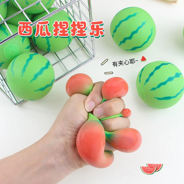A hand is squeezing a green watermelon-shaped stress relief toy, with other similar toys visible in a wire basket.