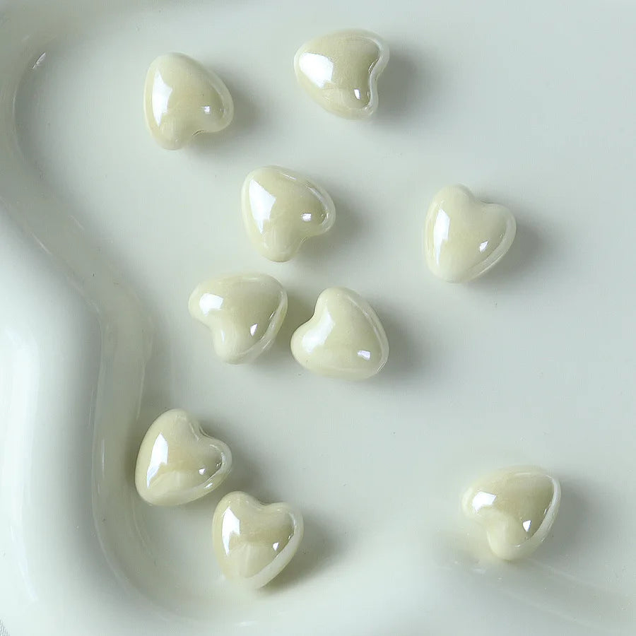 A collection of ten glossy, heart-shaped ceramic beads in an off-white or cream color arranged on a white surface.
