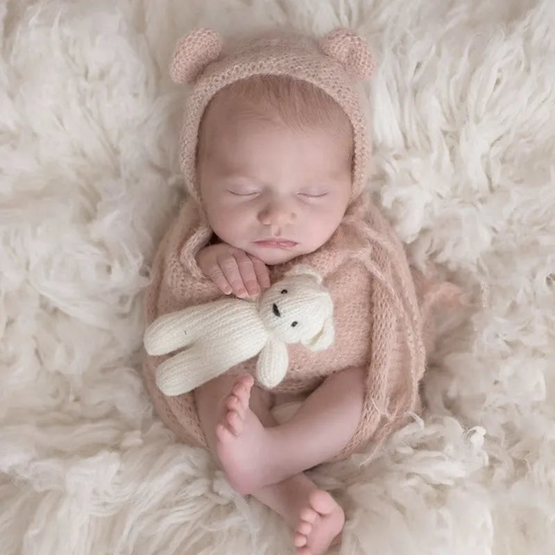A newborn baby sleeping wearing a knitted pink bear hat and matching wrap, resting on a fluffy white surface.