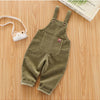 Olive green baby cotton overalls with adjustable straps lying on a wooden surface next to a camera.