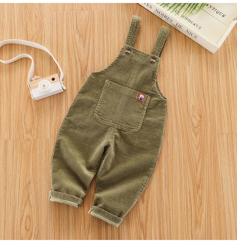 Olive green baby cotton overalls with adjustable straps lying on a wooden surface next to a camera.