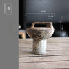 A vintage handcrafted ceramic vase with a textured, earthy brown color is shown on a wooden surface.