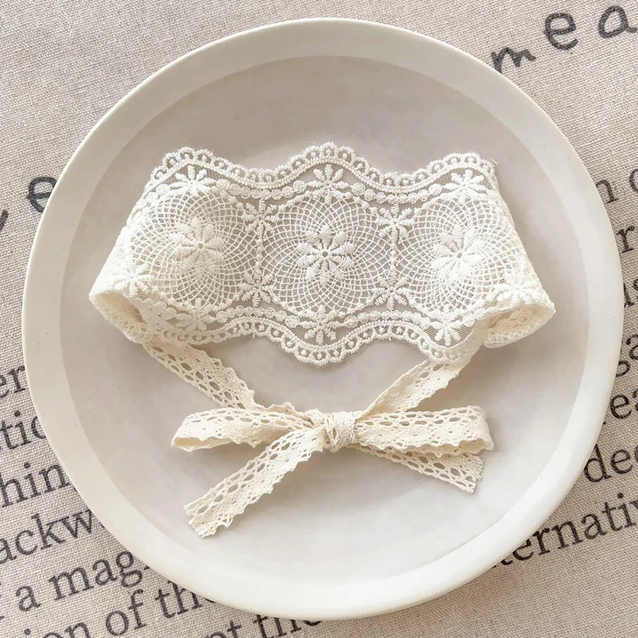 A white floral lace baby headband for newborns is displayed on a light ceramic plate.