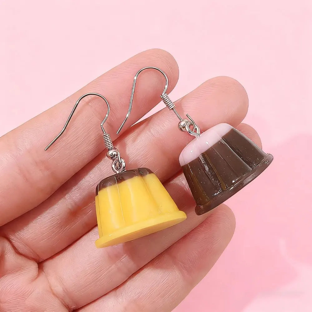 A hand holds a silver earring hook with a yellow pudding-shaped resin pendant and a white bead.