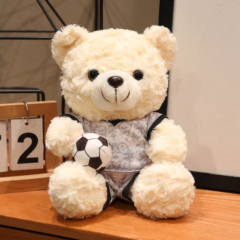 A fluffy cream-colored bear plush toy wearing a gray football patterned outfit and holding a soccer ball.