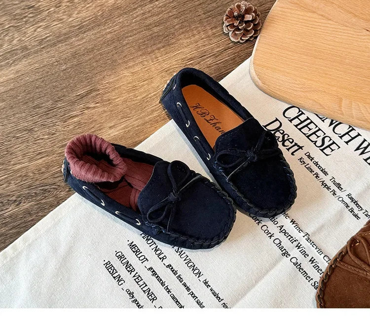 A pair of dark navy velvet slip-on moccasins with bow details resting on a white cloth.