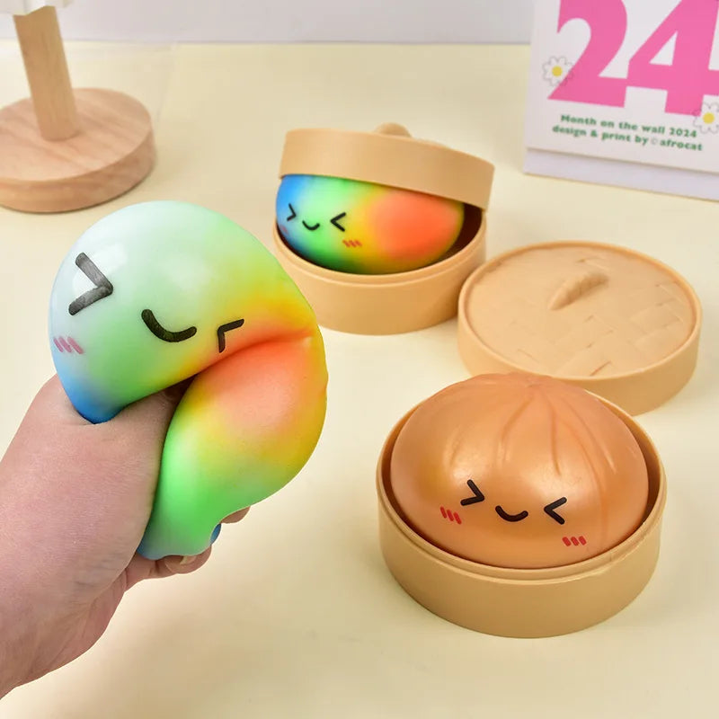 A hand holds a colorful, squishy dumpling stress relief toy with a smiling face in an open cardboard container.