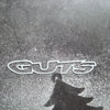 A silver vinyl sticker spelling out GUTS casts a dark shadow on a textured surface.