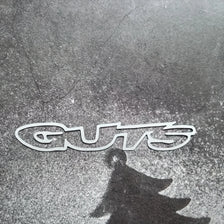 A silver vinyl sticker spelling out GUTS casts a dark shadow on a textured surface.