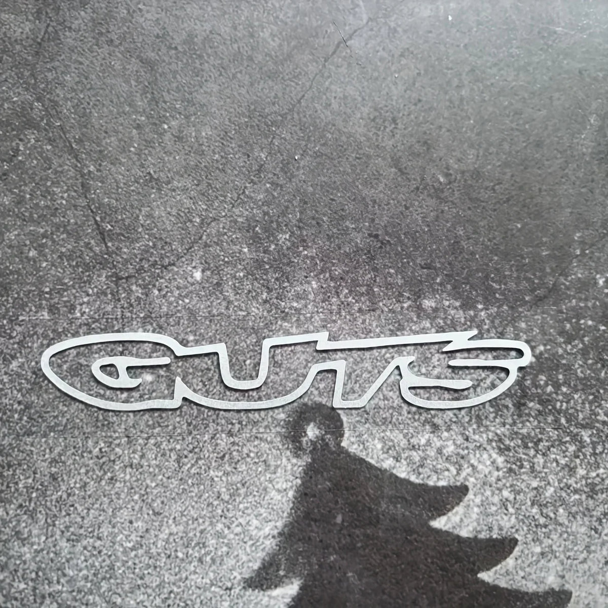 A silver vinyl sticker spelling out GUTS casts a dark shadow on a textured surface.