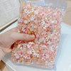 A hand holds a clear plastic bag filled with multicolor resin nail charms featuring pink and white floral designs.