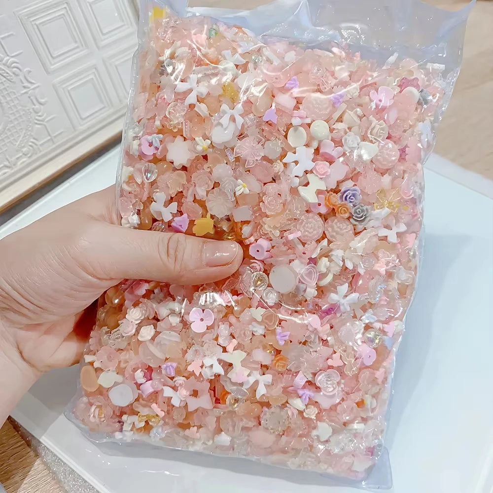 A hand holds a clear plastic bag filled with multicolor resin nail charms featuring pink and white floral designs.