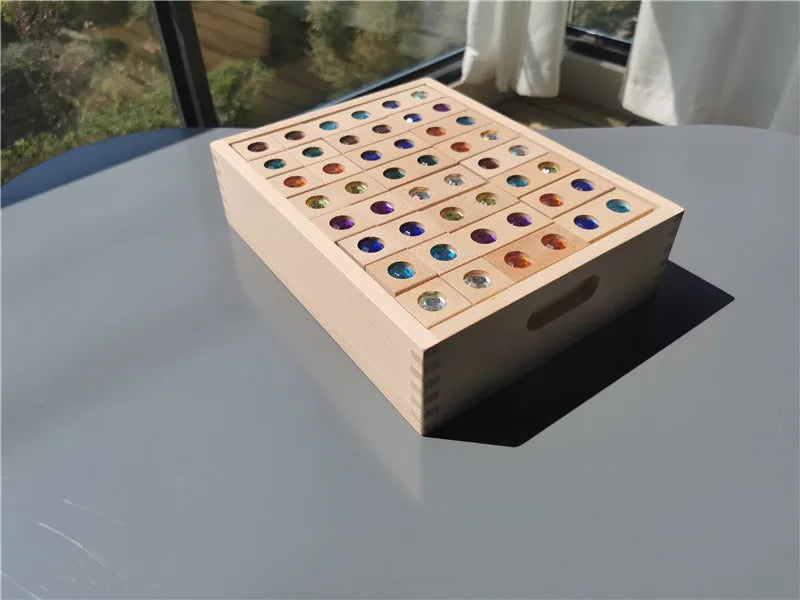 A wooden box filled with colorful acrylic gemstones arranged in a grid on a glass table.