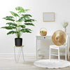 A large green faux monstera palm plant in a black pot sits on a white stand next to a white chair and a wooden desk.