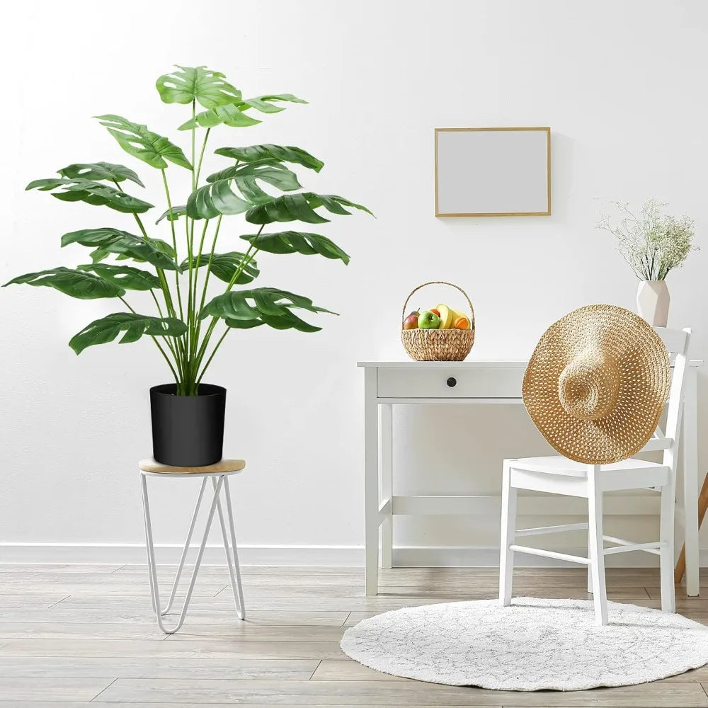 A large green faux monstera palm plant in a black pot sits on a white stand next to a white chair and a wooden desk.