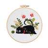 A white embroidery kit featuring a black cat and floral elements on a circular canvas with a wooden hanger.