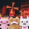 Three plush toys resembling cats in pink, orange, and white are displayed against a city backdrop.