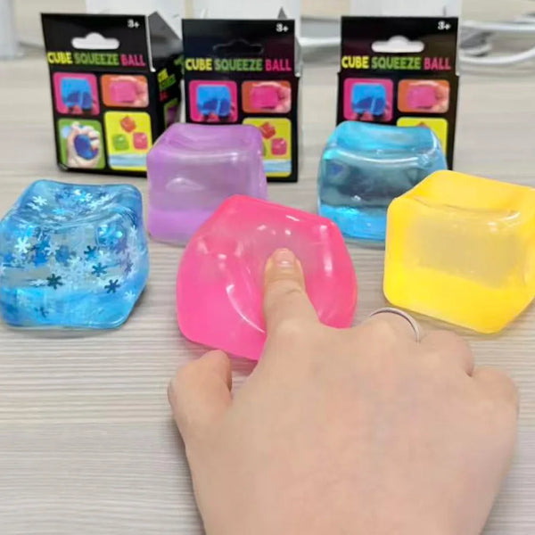 A hand is squeezing a bright pink cube squeeze ball toy, surrounded by various colored squishy balls.