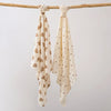 Two cream-colored baby gauze swaddle blankets with a delicate pattern are hanging on a wooden branch.