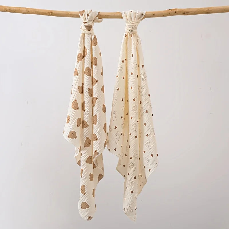Two cream-colored baby gauze swaddle blankets with a delicate pattern are hanging on a wooden branch.